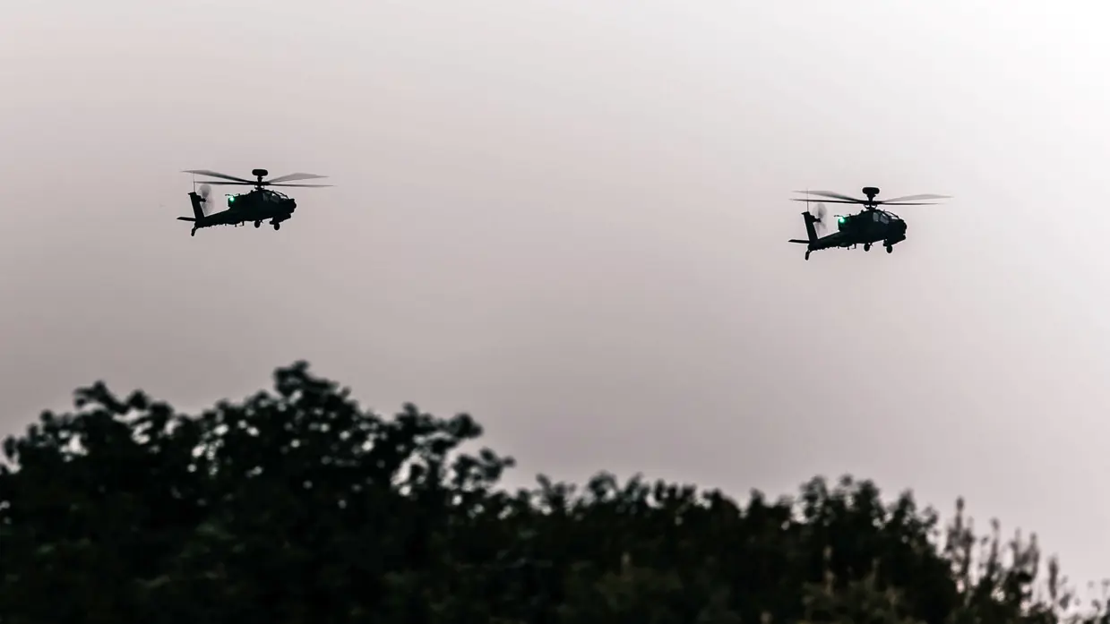 In de schemering vliegen twee Apaches met groene verlichting boven bomen.