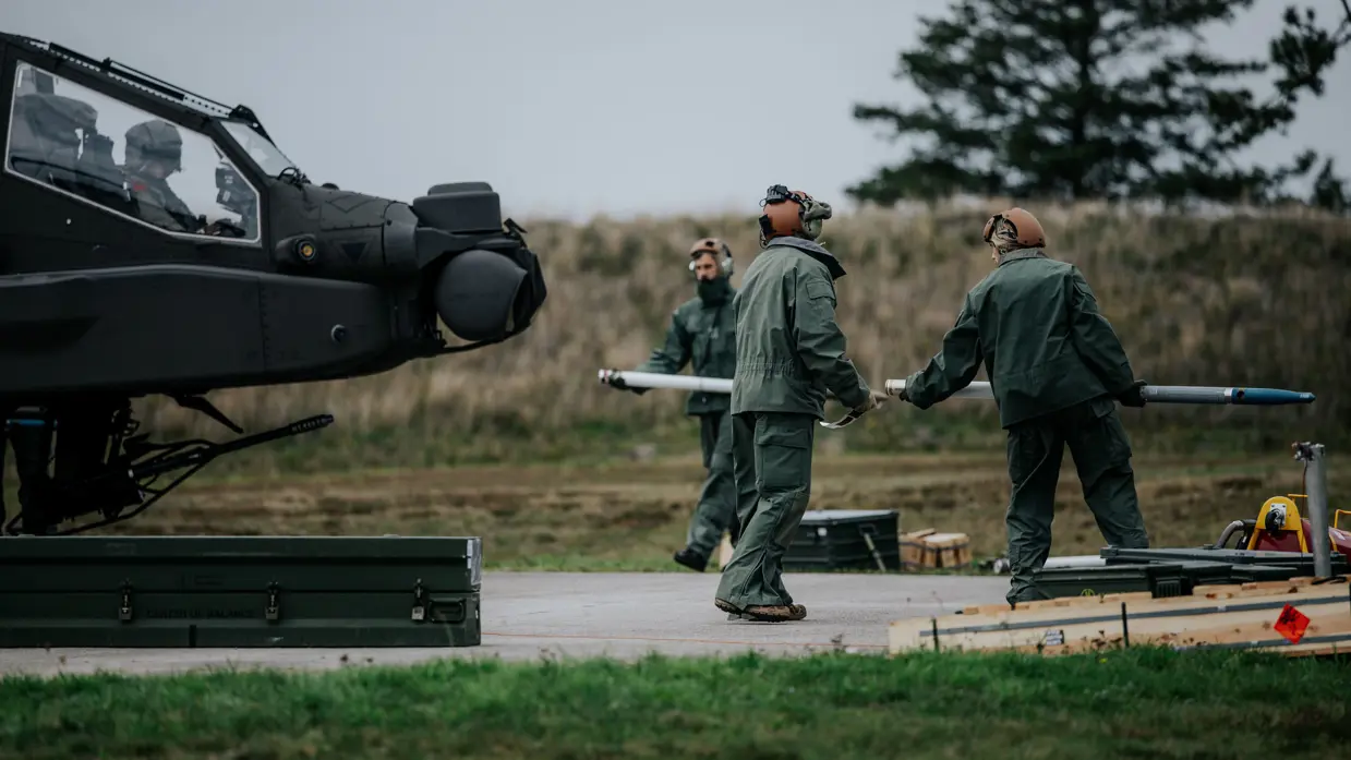 Drie personen in groene vliegerpakken en helmen dragen een lang, cilindervormig object , een raket, bij een donkere militaire helikopter. Grasveld met betonnen oppervlak, bomen op achtergrond.