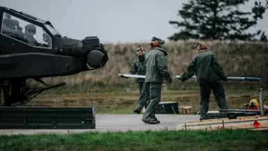 Drie personen in groene vliegerpakken en helmen dragen een lang, cilindervormig object , een raket, bij een donkere militaire helikopter. Grasveld met betonnen oppervlak, bomen op achtergrond.