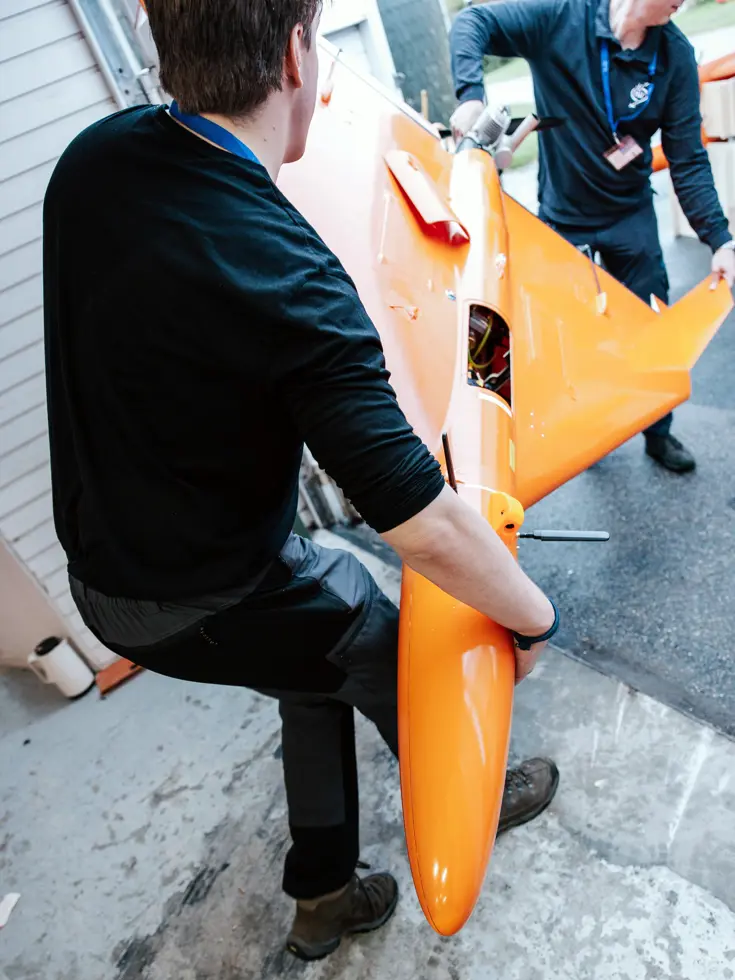 Twee mannen in donkere kleding draagt een grote oranje drone.