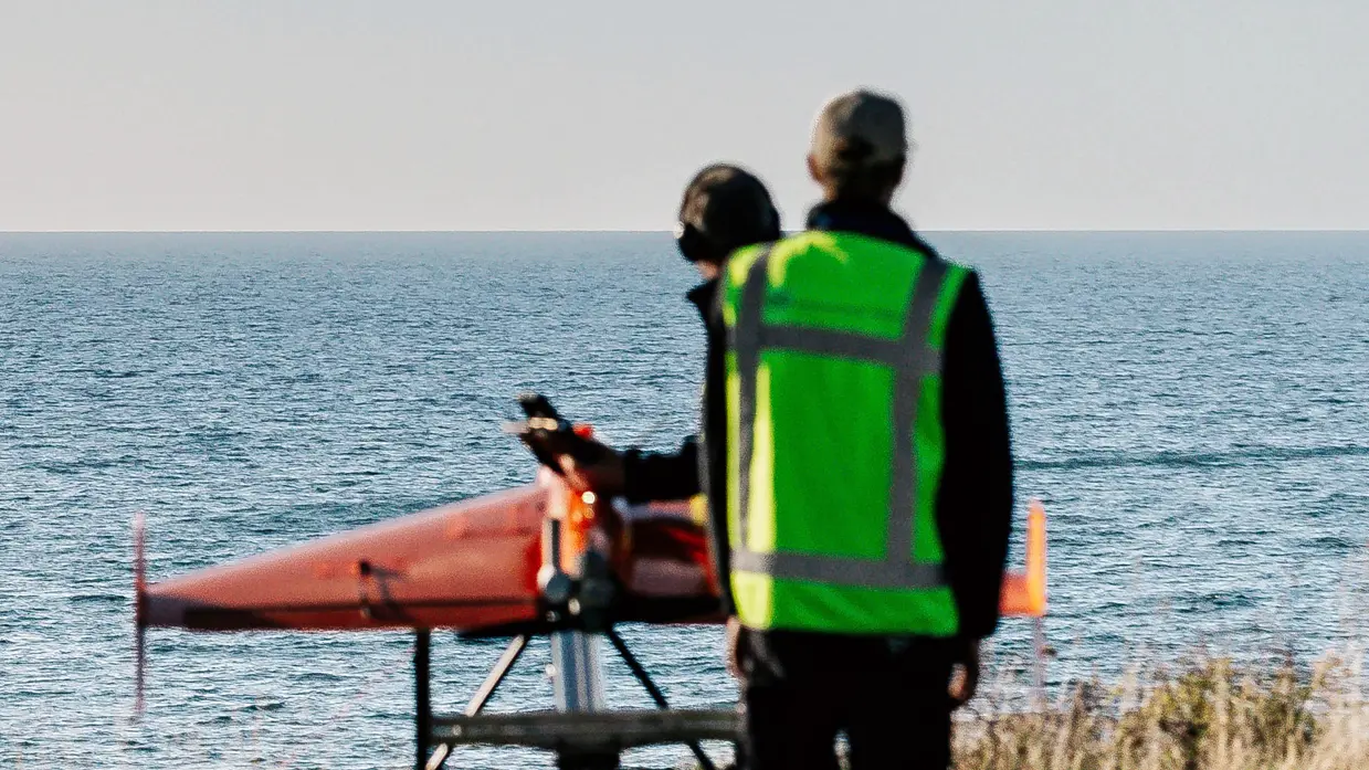 Een Apache hangt boven zee, met een oranje drone op een lanceerstelling daaronder.