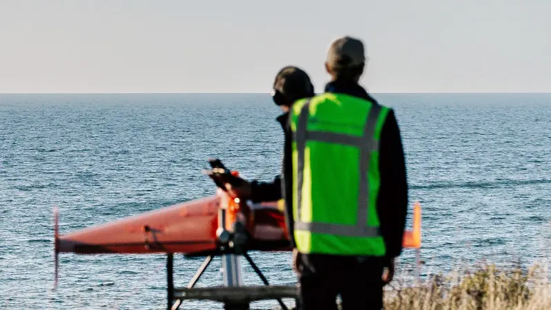 Een Apache hangt boven zee, met een oranje drone op een lanceerstelling daaronder.