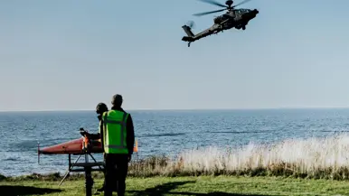 Een zwarte helikopter hangt boven water, op de grond staan twee mannen en een oranje gekleurd vliegtuigje. 