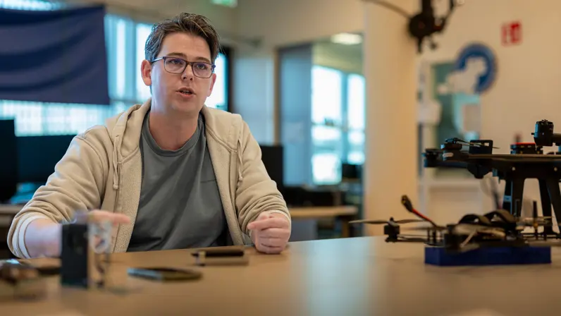Een bebrilde jongeman met hoodie en T-shirt aan legt iets uit aan een tafel vol onderdelen van een drone.