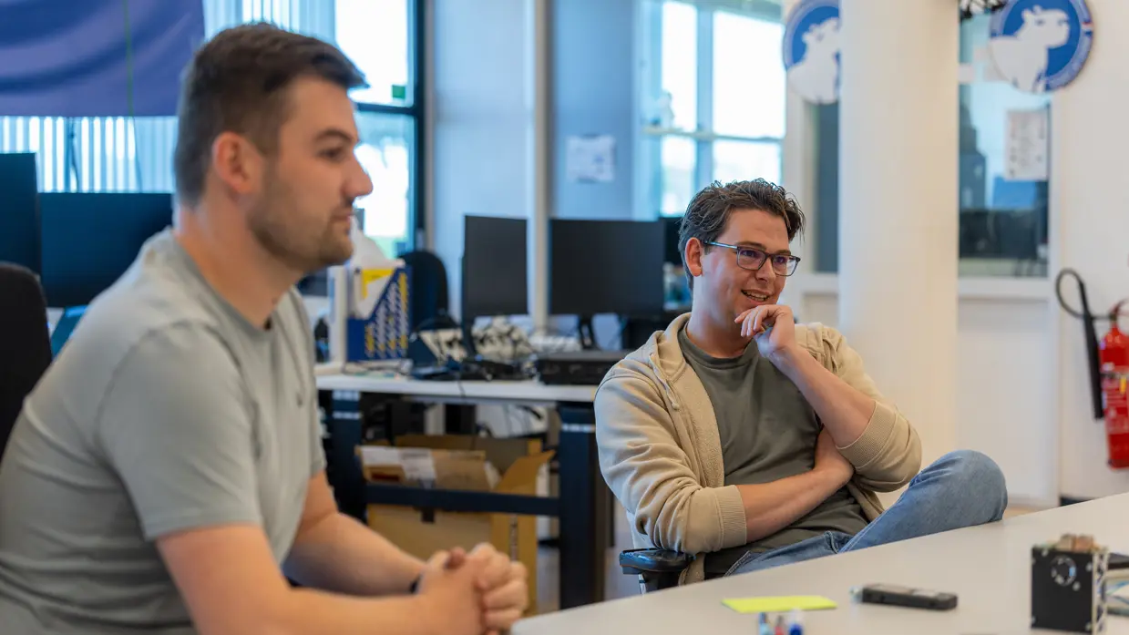  Jongeman in grijs shirt is met een bebrilde collega in gesprek aan een tafel vol drone-onderdelen.