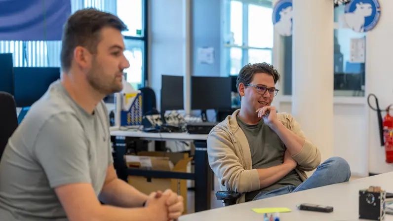  Jongeman in grijs shirt is met een bebrilde collega in gesprek aan een tafel vol drone-onderdelen.