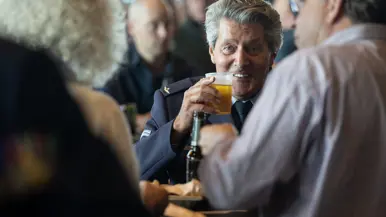 Een man met een biertje in zijn hand kijkt lachend de camera in.