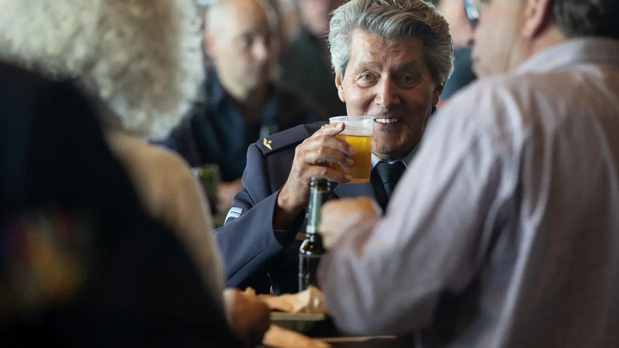 Een man met een biertje in zijn hand kijkt lachend de camera in.