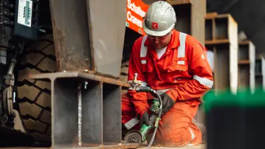 Een man in oranje pak met witte helm is bezig met slijpwerkzaamheden. Daarbij spatten de vonken ervan af. 