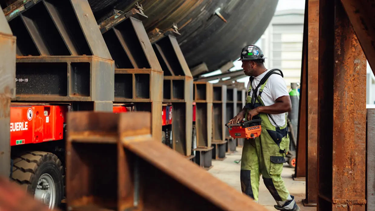 Een man met een groene overall bestuurt de knoppen op een oranje kastje, dat om zijn middel hangt. Hij loopt naast de constructie van de onderzeeboot.