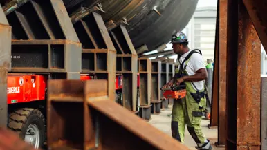 Een man met een groene overall bestuurt de knoppen op een oranje kastje, dat om zijn middel hangt. Hij loopt naast de constructie van de onderzeeboot.