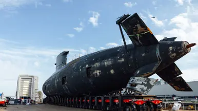 De onderzeeboot vanaf achteren gezien. Het vaartuig wordt verplaatst op kleine aaneengesloten karretjes. Een helikopter vliegt over.