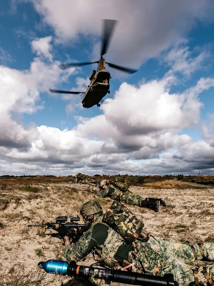 Op de grond liggen zwaar bewapende militairen met helmen op en een rugzak op de rug, terwijl een Chinook boven hun hoofd wegvliegt.