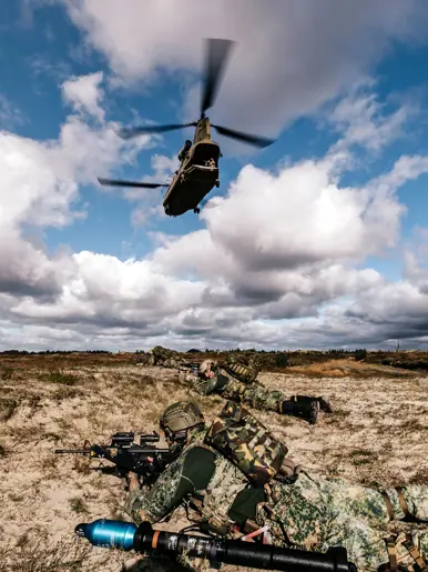 Op de grond liggen zwaar bewapende militairen met helmen op en een rugzak op de rug, terwijl een Chinook boven hun hoofd wegvliegt.