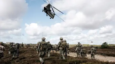 Een grote groep militairen met wapens en helm op lopen over een heidegebied. Een Chinook vliegt boven hen, in de verte vliegt er nog een.