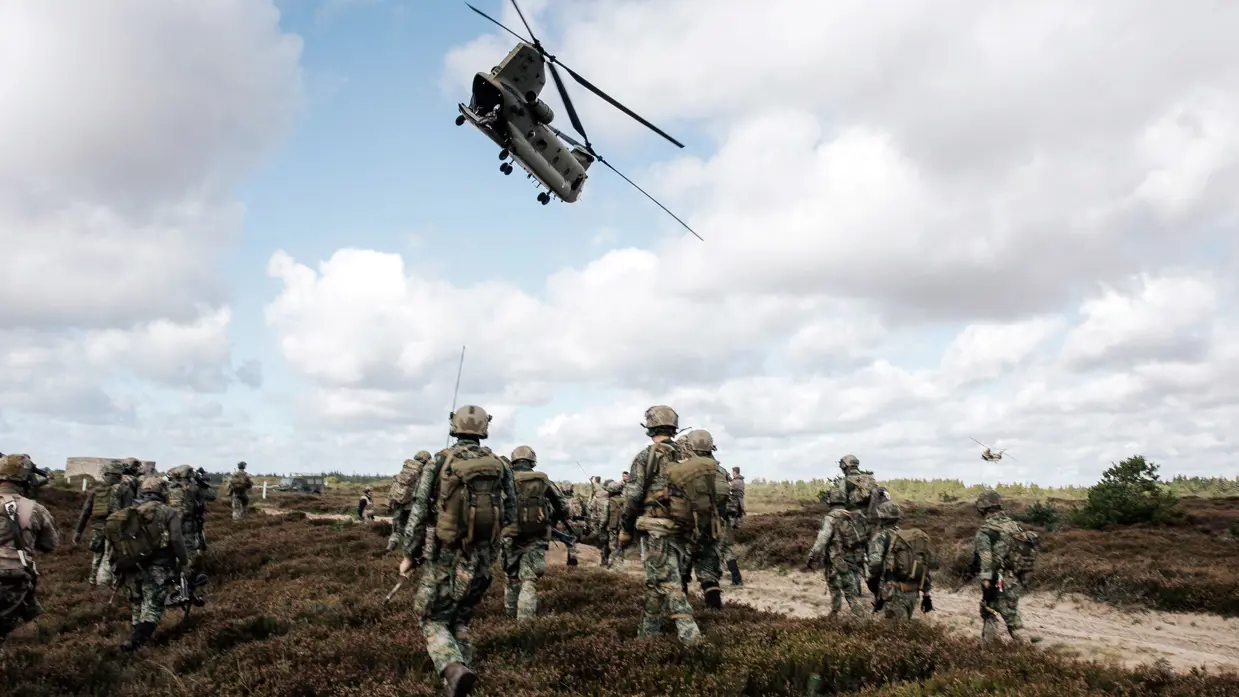 Een grote groep militairen met wapens en helm op lopen over een heidegebied. Een Chinook vliegt boven hen, in de verte vliegt er nog een.