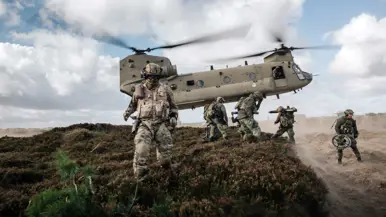 Een Chinook hangt vlak boven een heuvel. Militairen met helmen op lopen in de voorgrond.