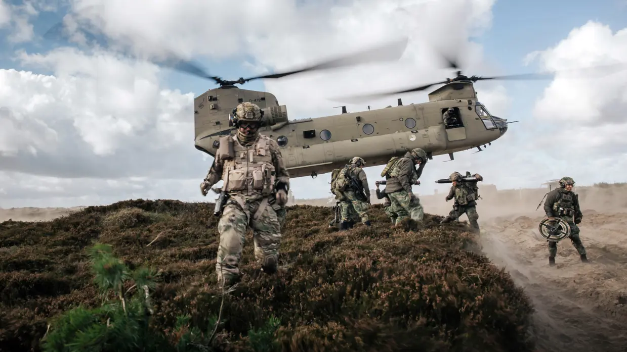 Een Chinook hangt vlak boven een heuvel. Militairen met helmen op lopen in de voorgrond.