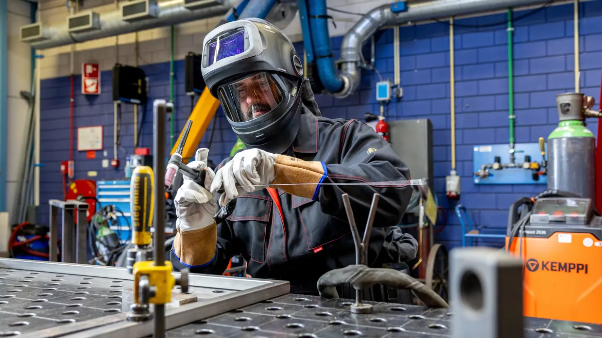 Erik met een lashelm en witte beschermhandschoenen is bezig met een lasverbinding. Hij houdt een lasdraad vast bij een metalen werkstuk op een werktafel met montagegaten.