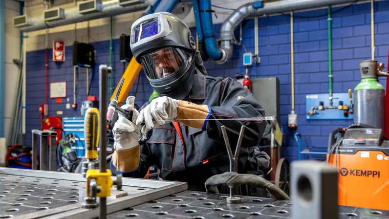 Erik met een lashelm en witte beschermhandschoenen is bezig met een lasverbinding. Hij houdt een lasdraad vast bij een metalen werkstuk op een werktafel met montagegaten.