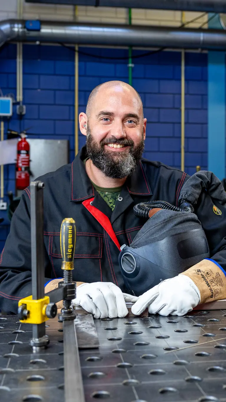 Sergeant 1 Erik van den Bergh poseert in lasuitrusting aan een metalen werkbank in een industriële werkplaats.