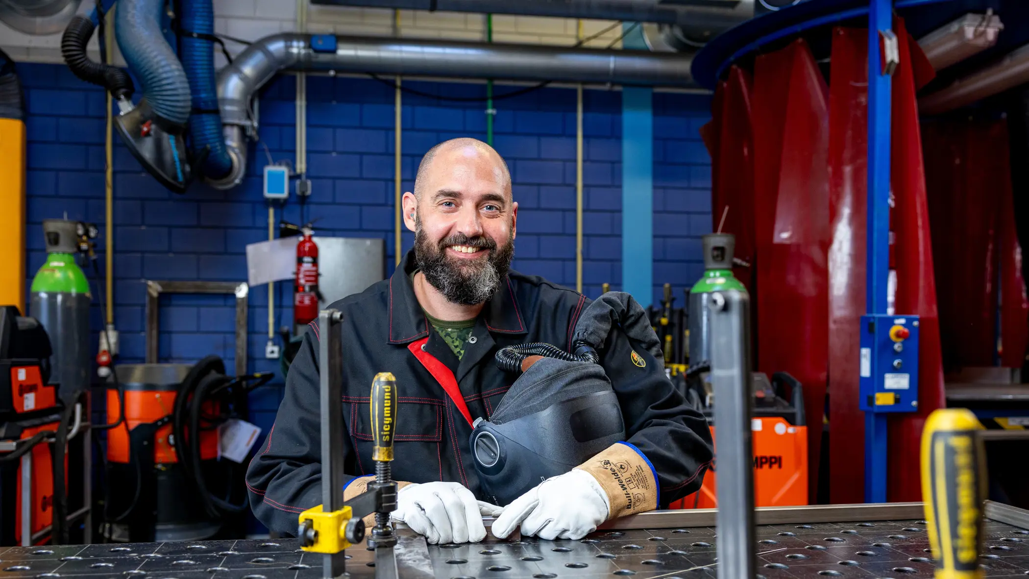 Sergeant 1 Erik van den Bergh poseert in lasuitrusting aan een metalen werkbank in een industriële werkplaats.