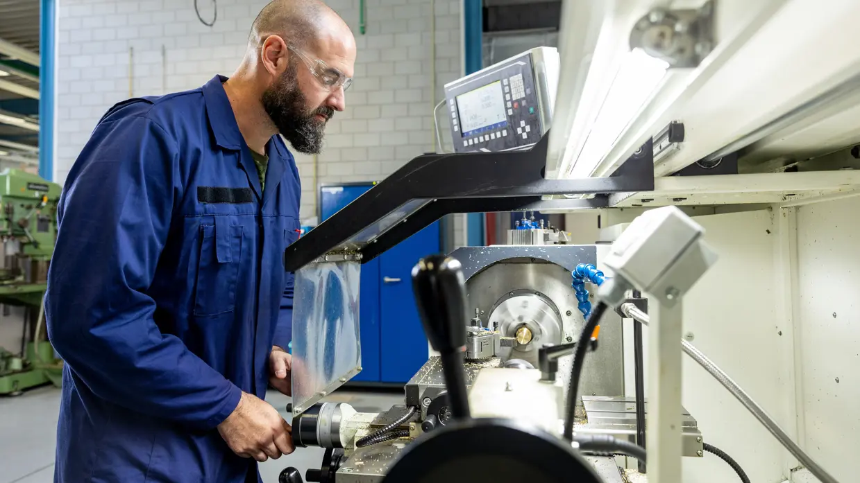 Erik met bril en in een blauwe werkoverall bedient een grote, witte industriële draaibank in een werkplaats.