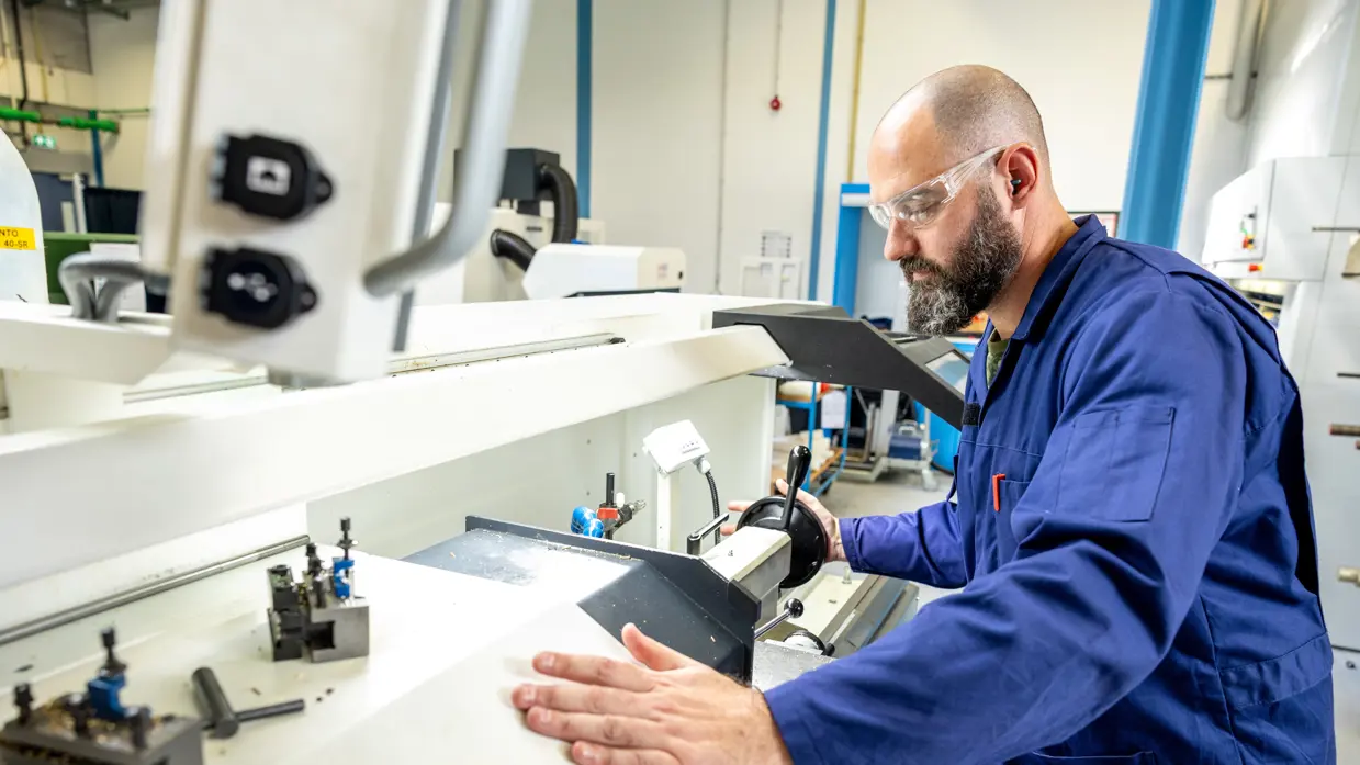 Erik met bril en in een blauwe werkoverall bedient een grote, witte industriële machine in een lichte werkplaats. Hij houdt zijn hand op een deel van de machine terwijl hij een hendel bedient.