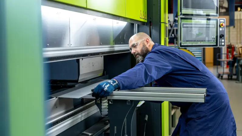 Erik in een blauwe werkoverall staat voor een grote, felgroene industriële machine. Hij leunt voorover en houdt met blauwe handschoenen een metalen plaat vast terwijl hij deze door de machine geleidt.