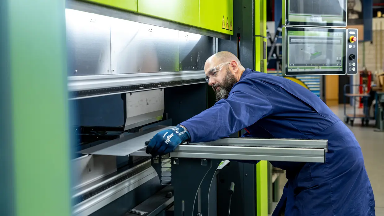 Erik in een blauwe werkoverall staat voor een grote, felgroene industriële machine. Hij leunt voorover en houdt met blauwe handschoenen een metalen plaat vast terwijl hij deze door de machine geleidt.