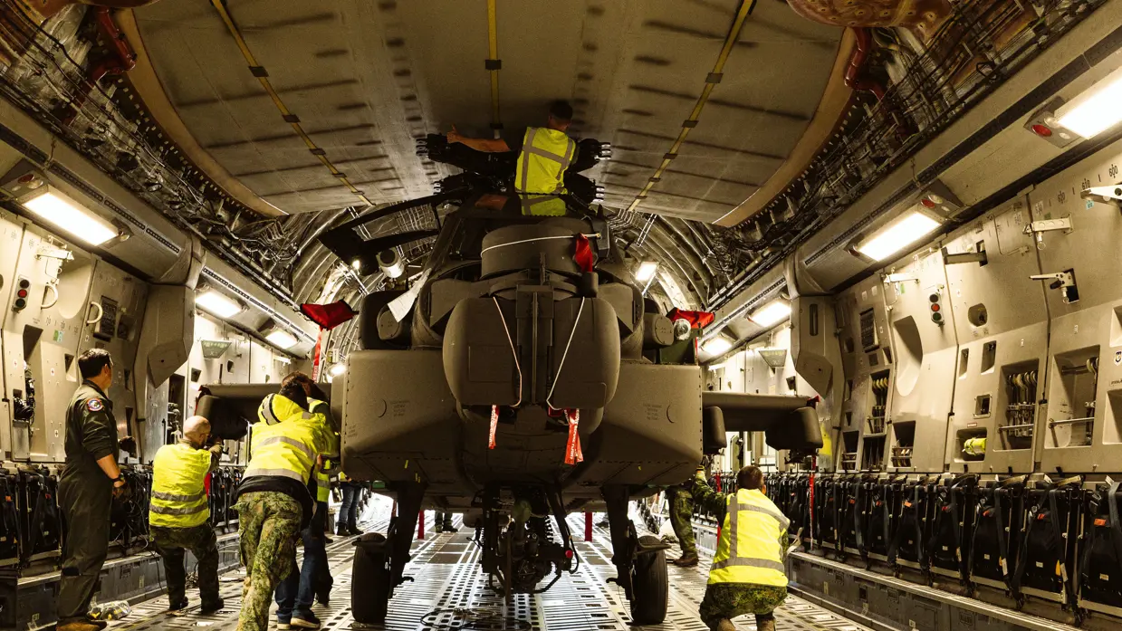 Militairen in gele hesjes staan in een laadruim en duwen een Apache naar buiten.