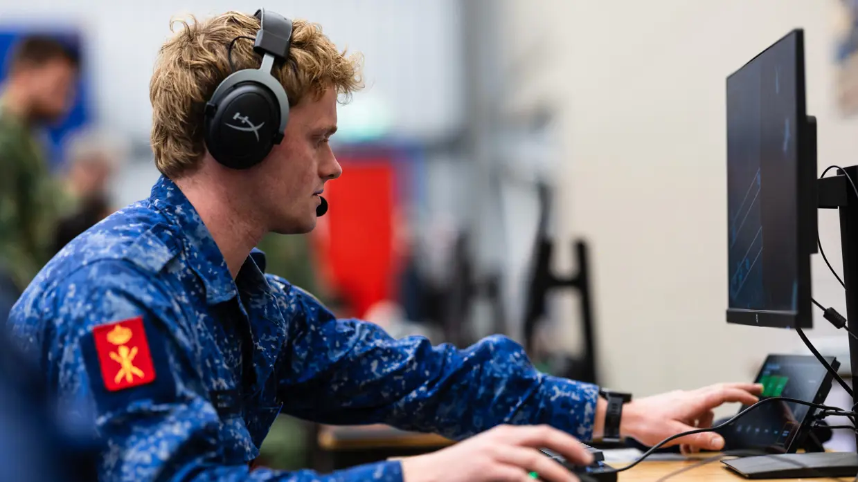 Een militair in opleiding zit met een koptelefoon op achter een computer