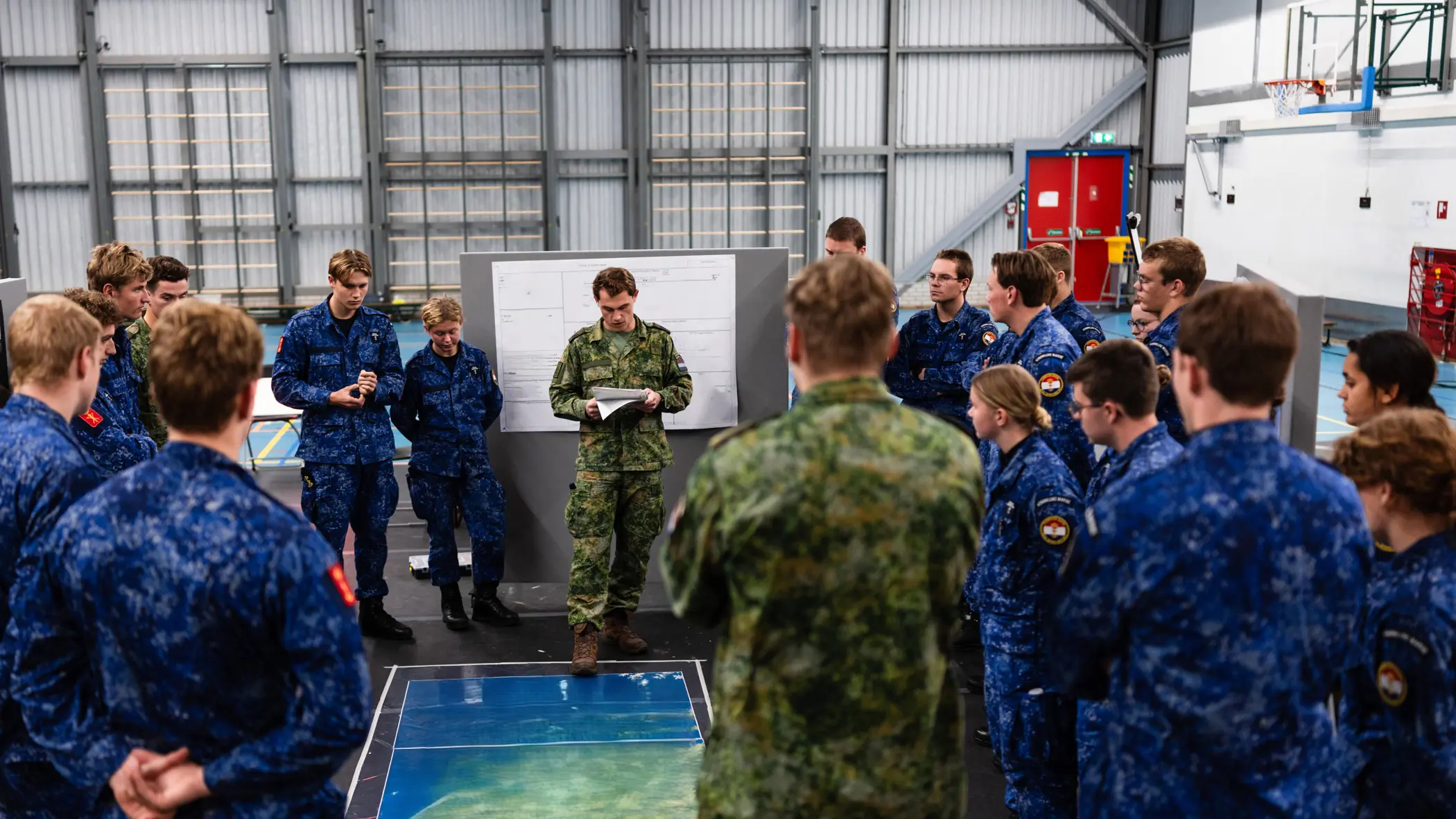Militairen van de marine in opleiding luisteren in een zaal naar een andere militair. Op de grond ligt een landkaart.