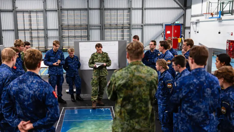 Militairen van de marine in opleiding luisteren in een zaal naar een andere militair. Op de grond ligt een landkaart.