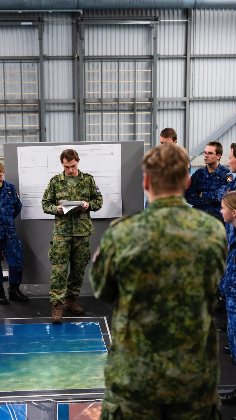 Militairen van de marine in opleiding luisteren in een zaal naar een andere militair. Op de grond ligt een landkaart.