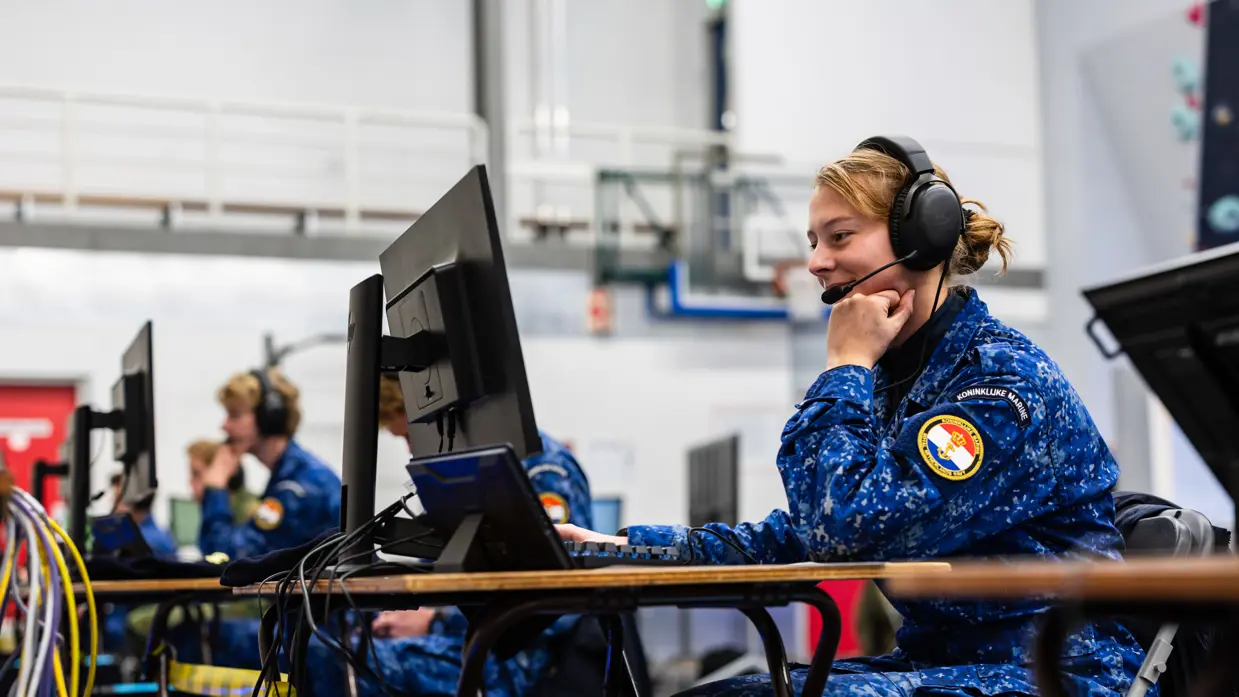 Een vrouwelijke militair zit met een koptelefoon op achter een computer.