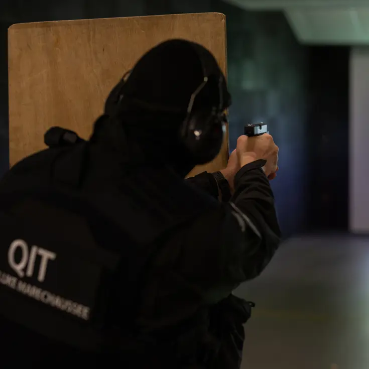 Een marechaussee richt zijn wapen vanachter een houten schot op het doel verderop.
