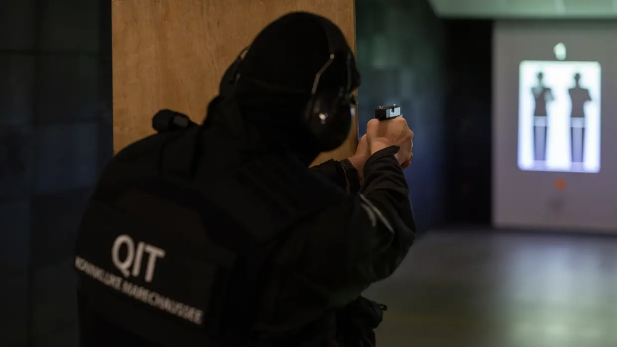 Een marechaussee richt zijn wapen vanachter een houten schot op het doel verderop.