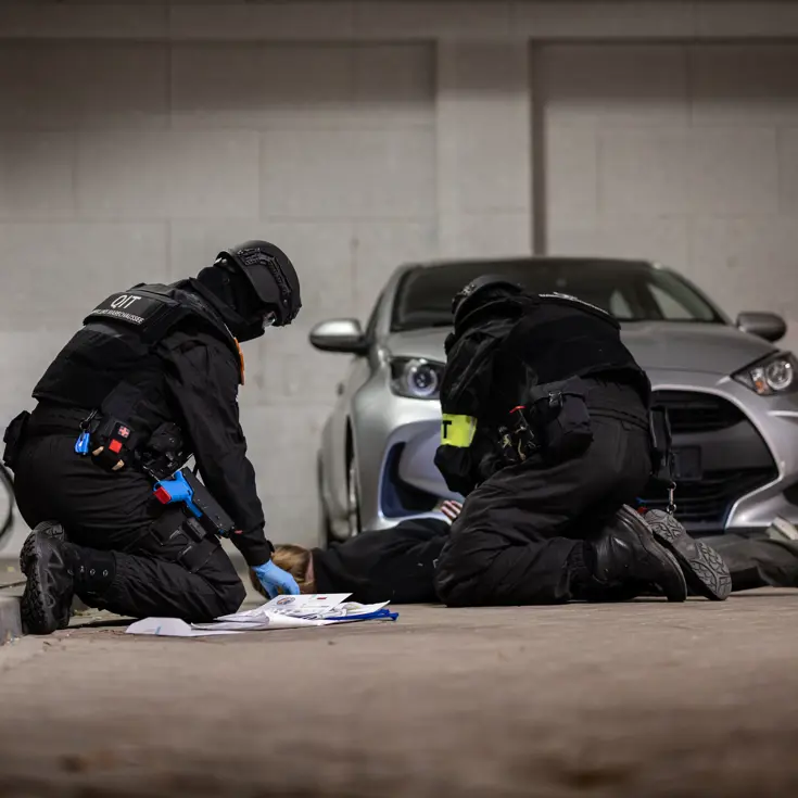 Twee marechaussees knielen bij een geboeide verdachte die op de grond ligt.
