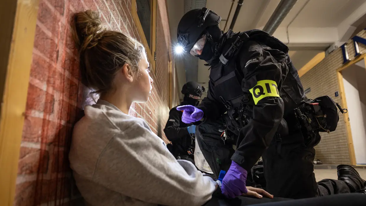 Een marechaussee knielt bij een gewonde dame en spreekt haar aan.