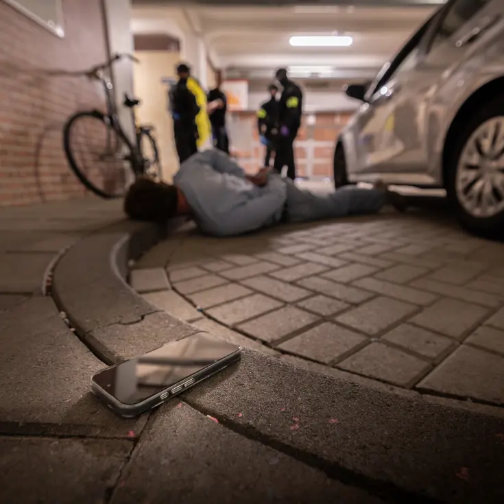 Een verdachte ligt op de grond met zijn hoofd op de stoep. Op de voorgrond een telefoon.