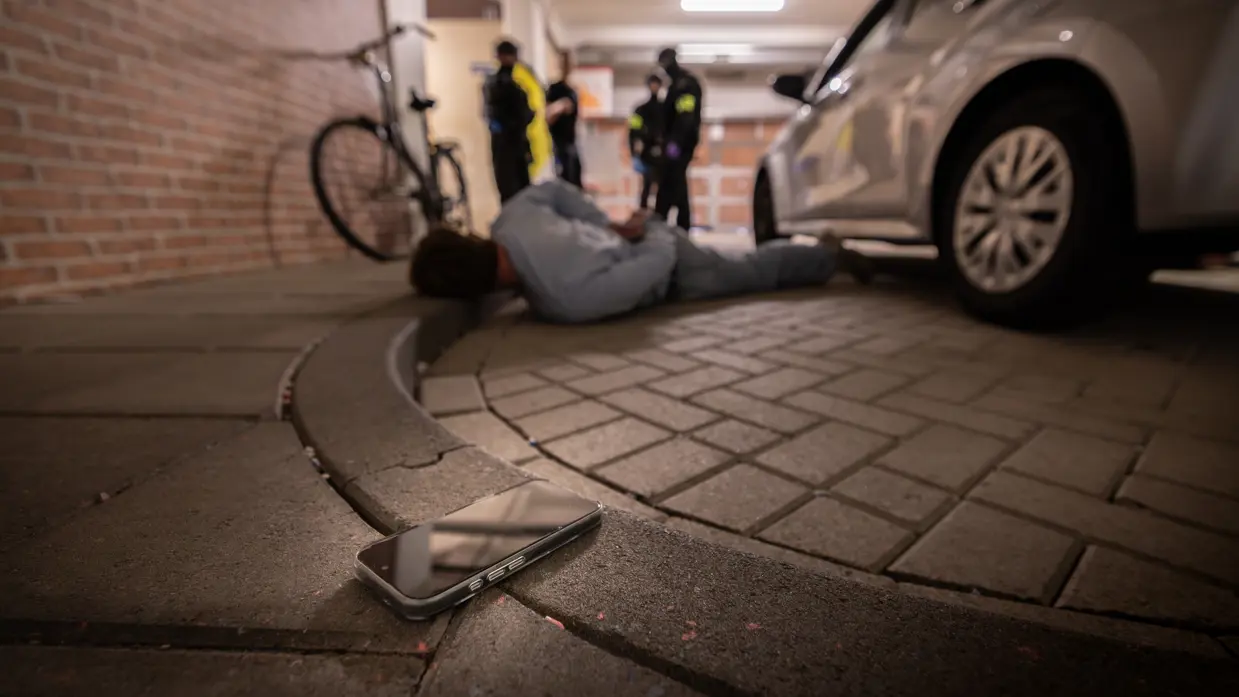 Een verdachte ligt op de grond met zijn hoofd op de stoep. Op de voorgrond een telefoon.