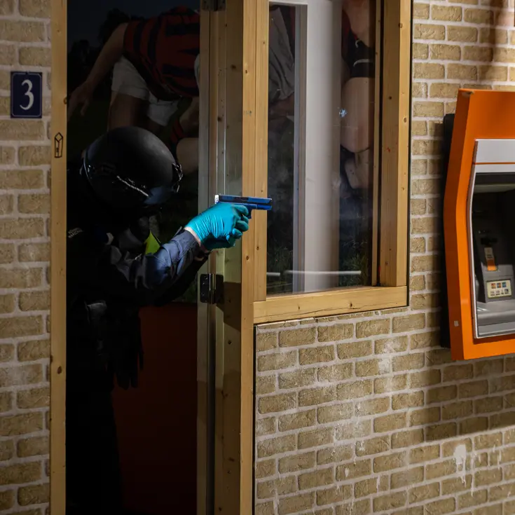 Een marechaussee met getrokken wapen kijkt om de hoek vanuit een deuropening.