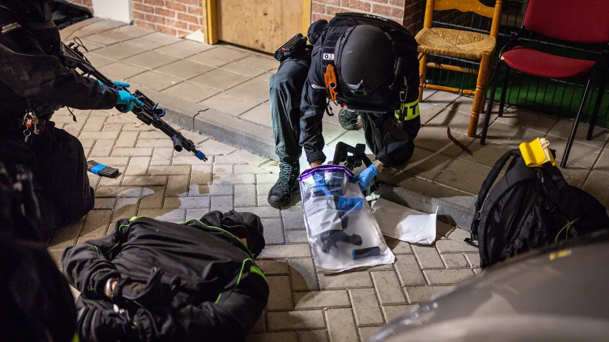 Terwijl de verachte op de grond ligt, stelt een marechaussee het wapen veilig.