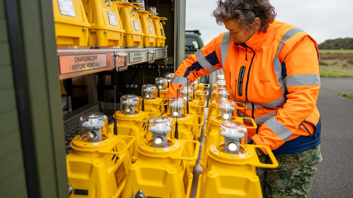Een vrouw in oranje jas haalt een lamp met een gele onderkant uit een groene aanhangwagen.