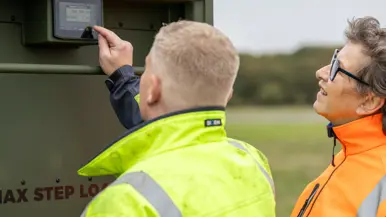 Een man in gele jas wijst naar een display op de groene aanhanger. Een vrouw in oranje jas met bril kijkt mee.  