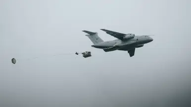 Uit een wit straalvliegtuig met een antenne op de neus valt een kist aan een parachute.