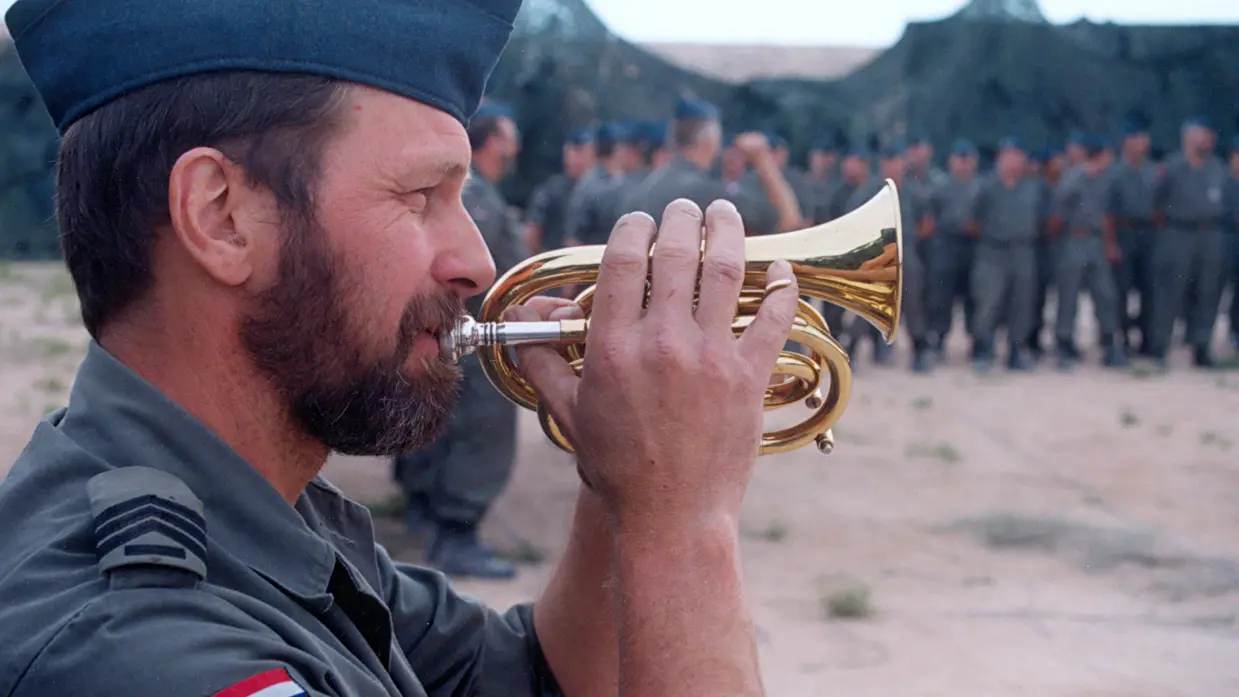 Een hoornblazer bespeelt zijn instrument tijdens het appèl. In de achtergrond staan militairen op een rij. Daarachter staan groene tenten.