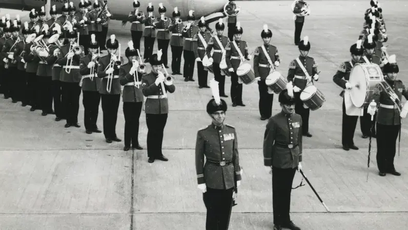 Foto op een vliegveld: een militaire kapel in uniform met hoge hoeden en witte pluimen staat in formatie met trompetten, trombones en drums; op de achtergrond wacht een groot vliegtuig.