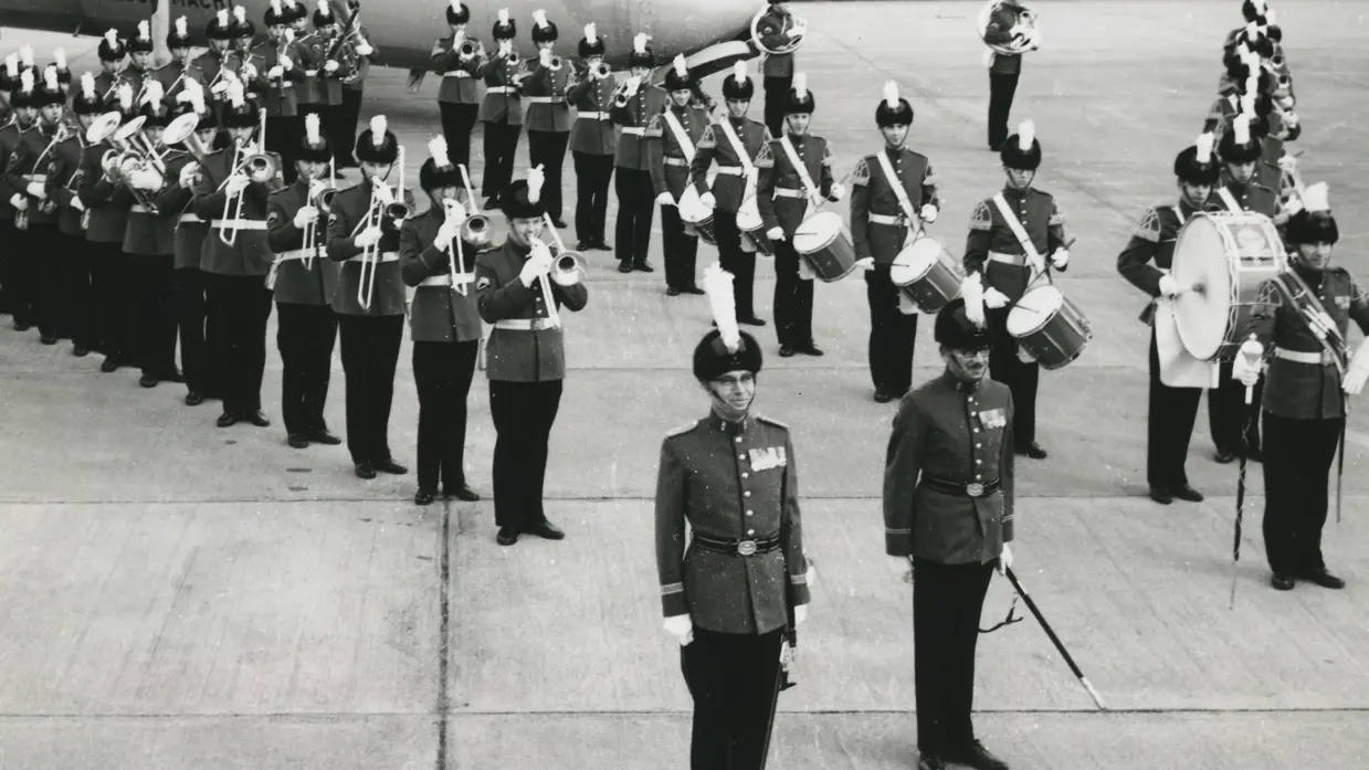 Foto op een vliegveld: een militaire kapel in uniform met hoge hoeden en witte pluimen staat in formatie met trompetten, trombones en drums; op de achtergrond wacht een groot vliegtuig.